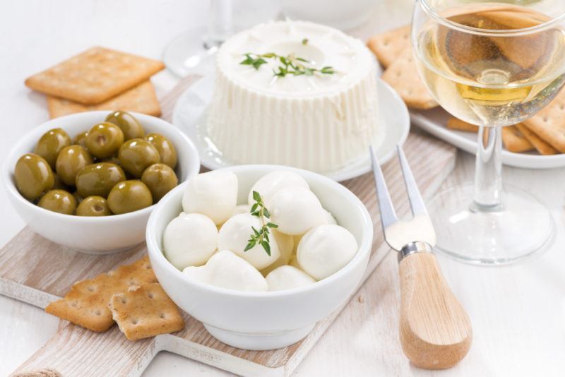 A bowl of fresh feta, green olives, and ricotta cheese, next to a glass of wine and a fork