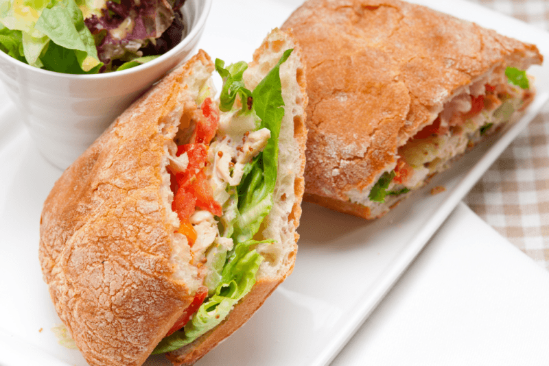 A large white plate with a fresh ciabatta sandwich with lettuce, tomato, and meat. The sandwich has been cut in half and there is a small dish of salad in the background.