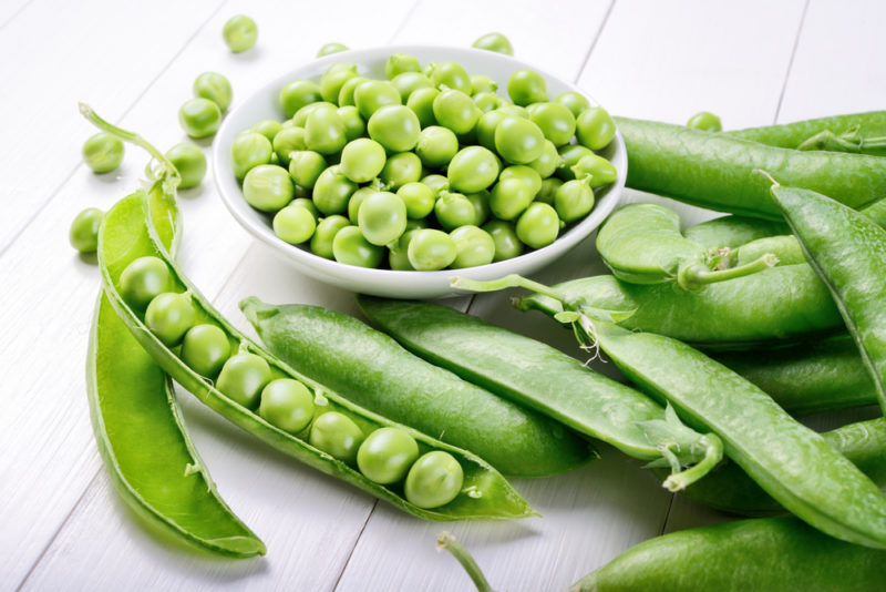 A white wooden table with various whole green pea pods, along with one opened pod and a white bowl of green peas