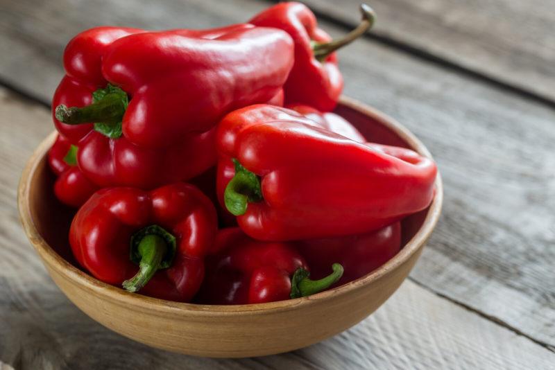 Five whole red bell peppers stacked on top of the other in a ceramic bowl