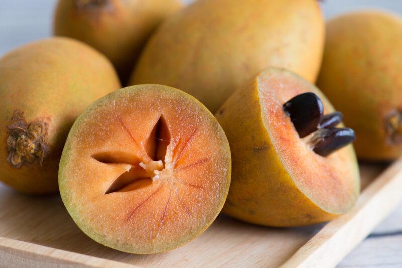 A wooden board with a collection of whole sapodilla fruits, along with one that has been cut in half