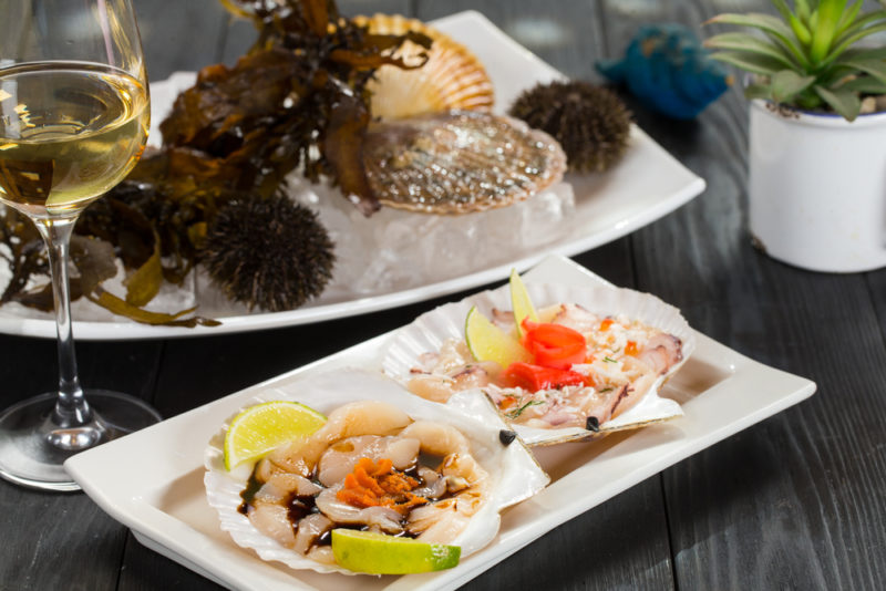 A white plate with scallops, next to a glass of white wine with sea urchins on another plate in the background