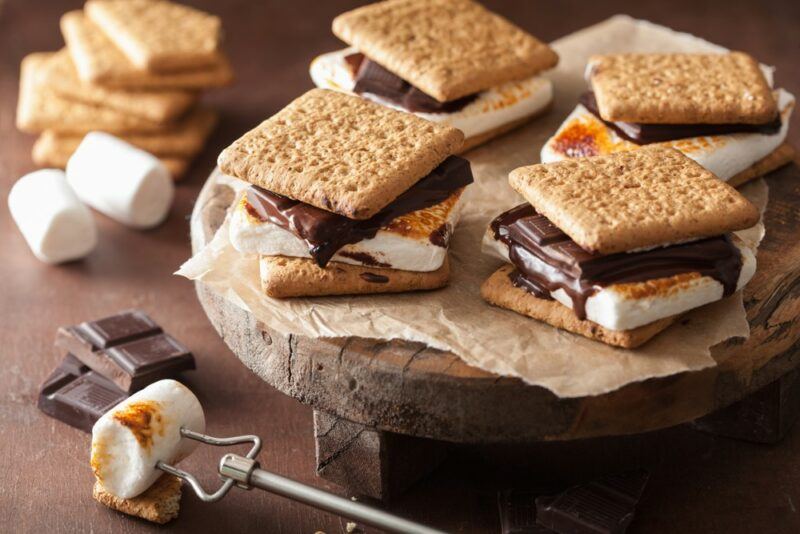 A wooden board with paper that has four freshly prepared s'mores on top, with more s'mores, marshmallows, and graham crackers next to it