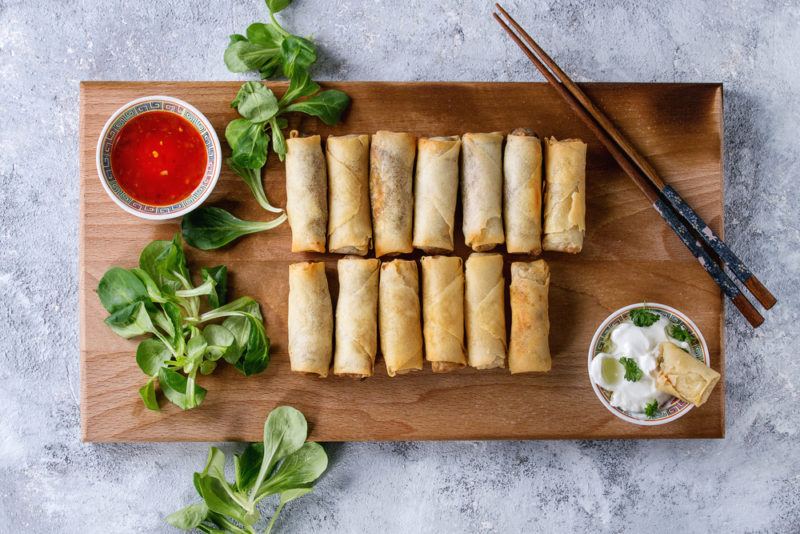 A wooden board with a dozen or so fried spring rolls with dips, greenery, and chopsticks