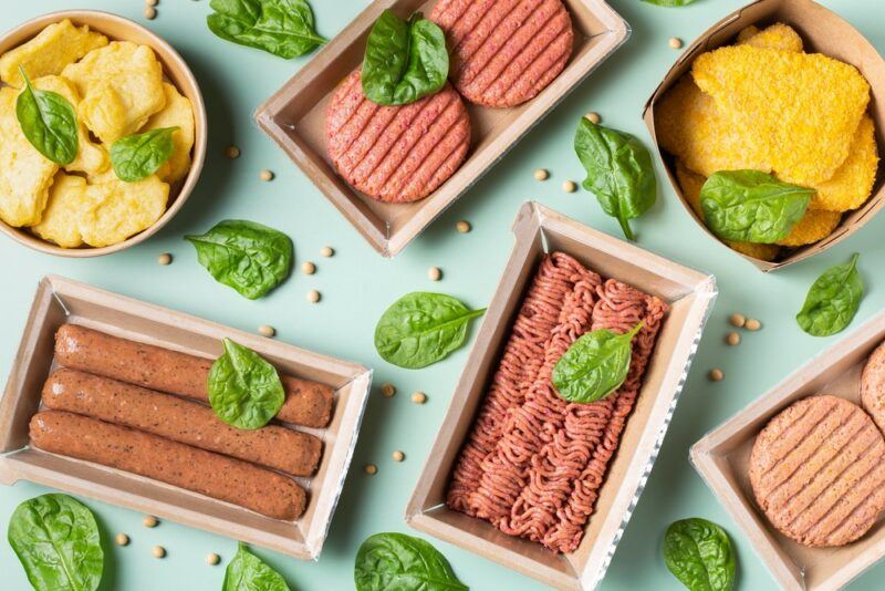 A light green table with various containers of raw plant-based meats, including sausages, mince, and chicken