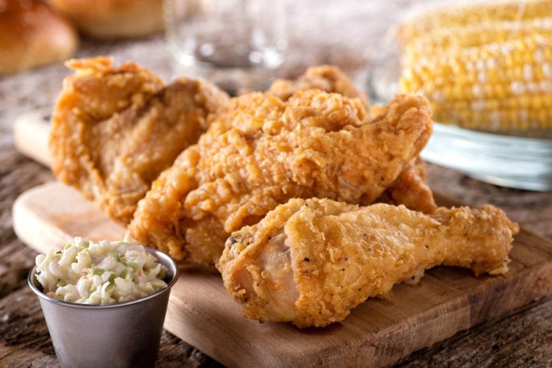 Fried chicken drumsticks on a wooden board near a small container of coleslaw and corn on the cob