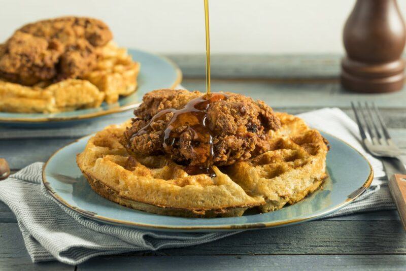 Two plates containing chicken and waffles, where one is being drizzled with maple syrup