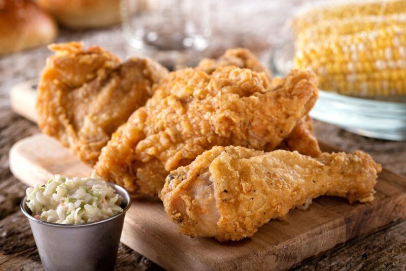 A wooden board containing fried chicken, next to some coleslaw and corn