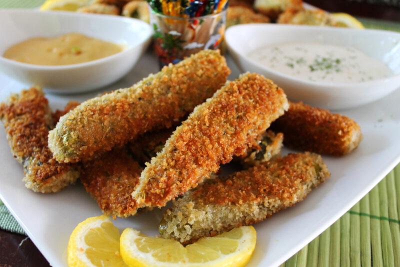 A white plate with some sliced lemons, two white containers of dip and various fried dill pickles