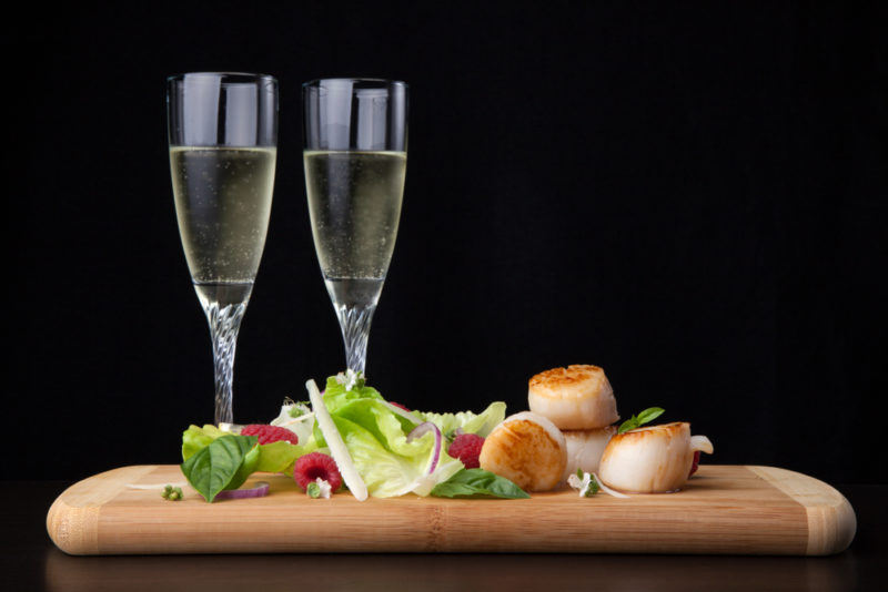 A wooden tray with some scallops and a salad, next to two glasses of chardonnay against a black background