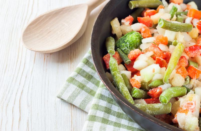 A black bowl of frozen vegetables on a table next to a wooden spoon