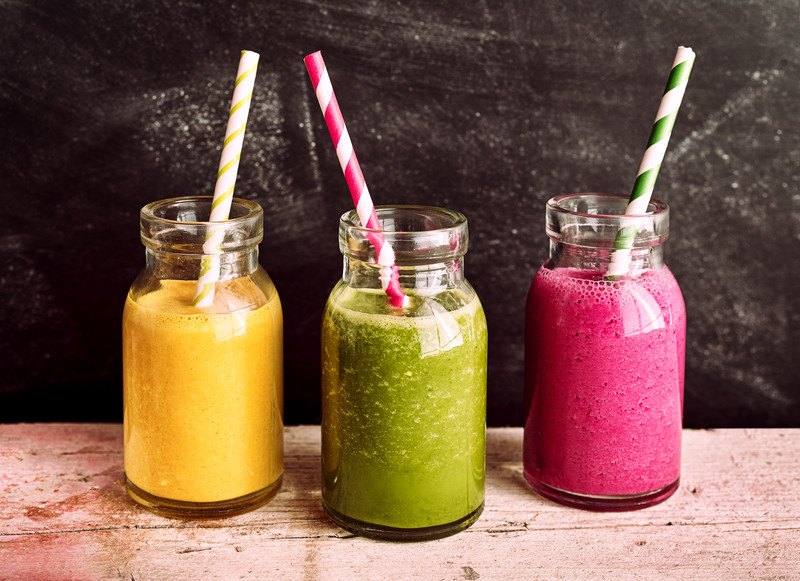 Three glass jars with pureed foods in orange, green, and purple with striped straws in them rest on a wooden board.