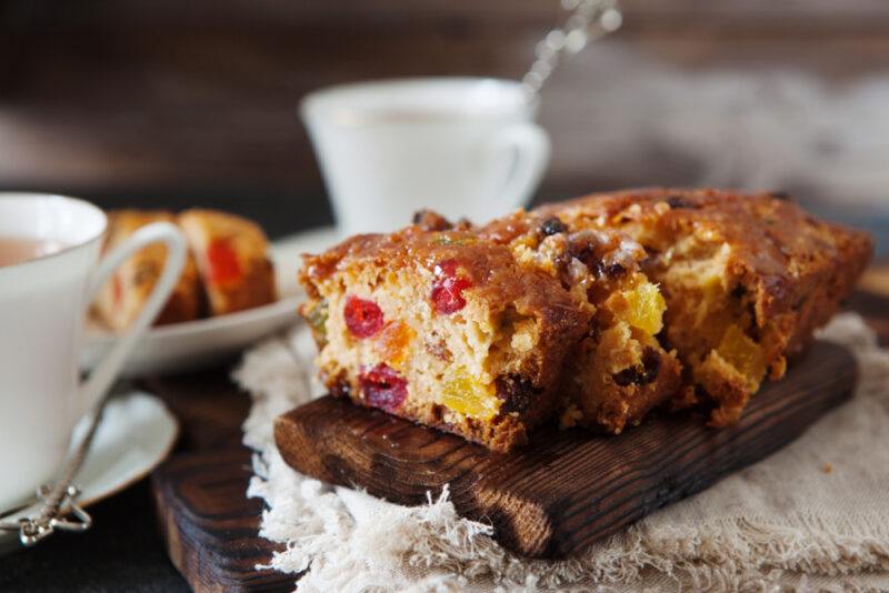 on a wooden plank are sliced fruit cake with a dish at the back with a sliced of fruit cake and cups of tea