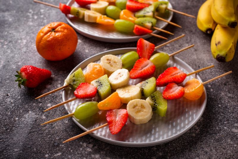 Two plates with fruit skewers and a couple of pieces of fruit on a table