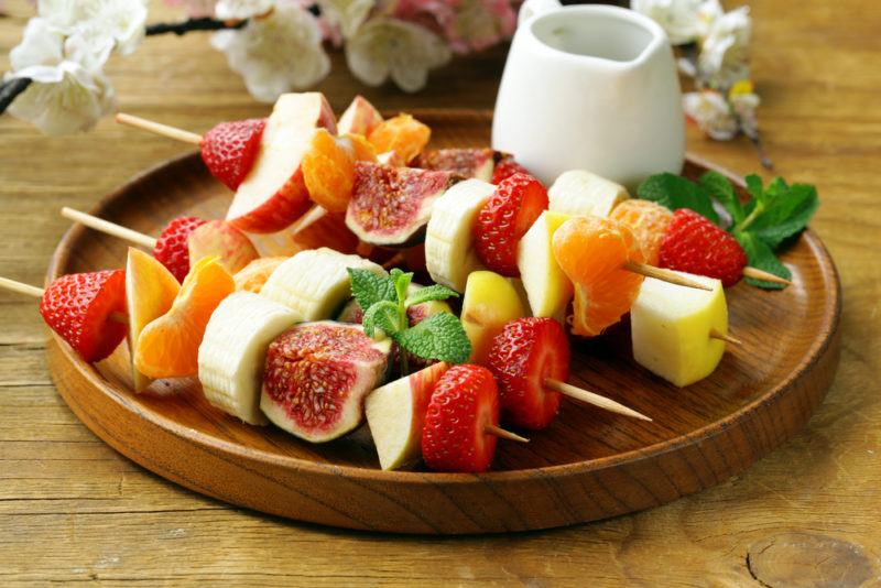 A wooden plate with skewers of fruit and cheese next to a glass of milk or some type of dip