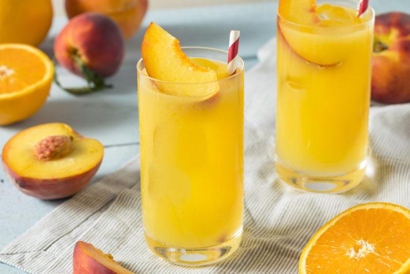 Two tall glasses containing a fuzzy navel cocktail, with peaches and oranges on a table