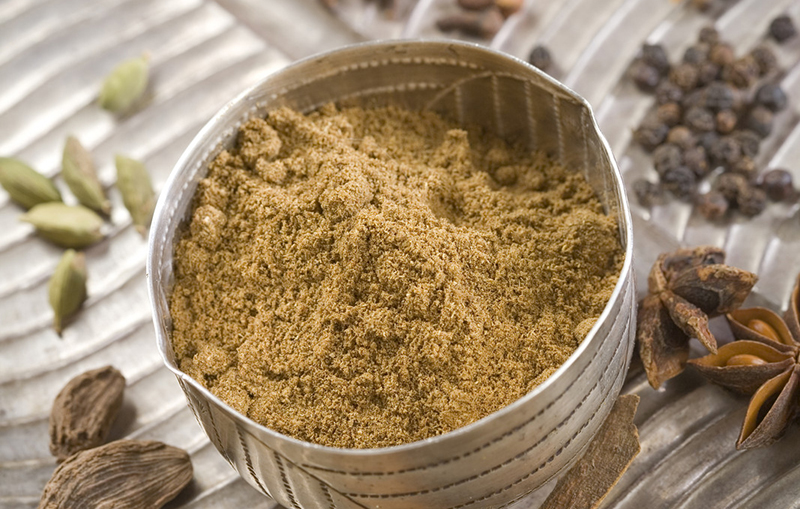 top view image of a metal bowl full of garam masala on a metal textured platter with star anise, cardamom, fennel, cloves, and black peppercorns