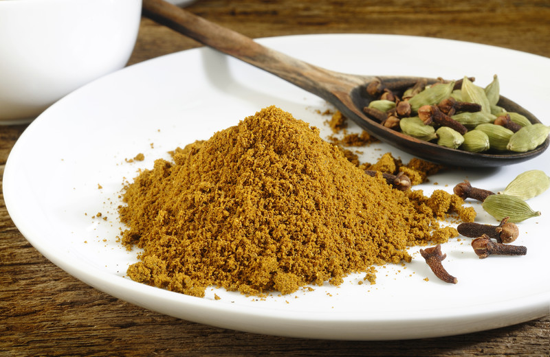 A mound of garam masala on a white ceramic plate with loose whole cloves and fennel seeds around it and a wooden spoon with whole cloves and fennel seeds.