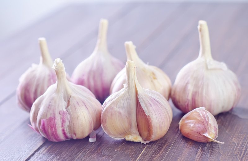 This photo shows six bulbs of garlic and one loose clove of garlic, all with a purple tint, sitting on a wooden table.