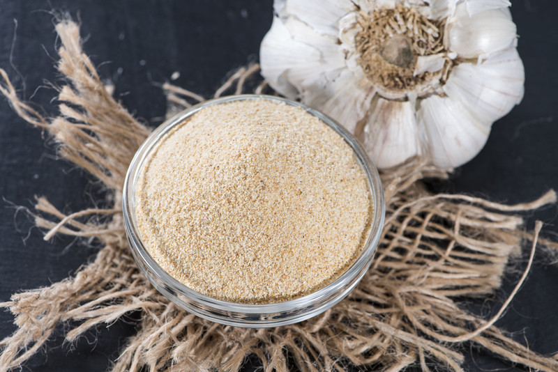 Small glass bowl of garlic powder resting on a piece of sack cloth on top of a black surface with a head of garlic at the back.