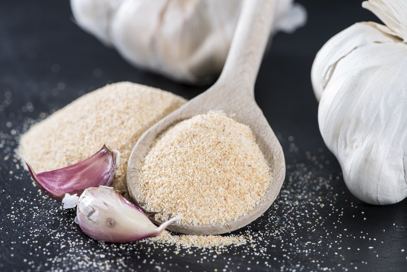 closeup image of a wooden spoonful of garlic powder with fresh garlic cloves and head beside it