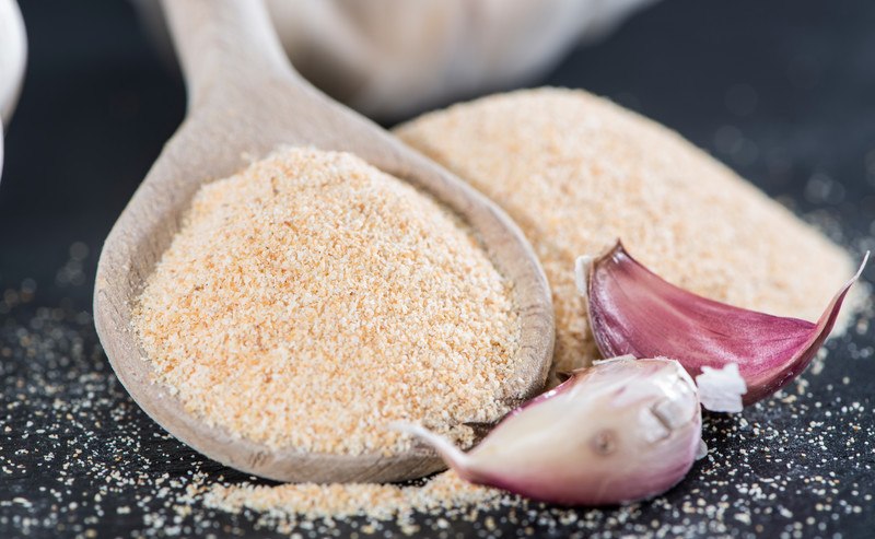 a wooden ladle full of garlic powder with a couple of garlic cloves beside it and a mound of garlic powder behind it