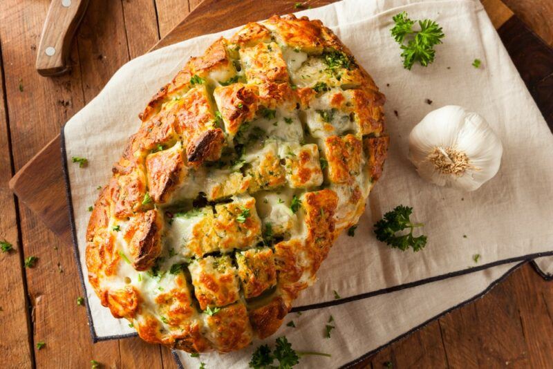 A wooden board containing a large piece of garlic and parsley pull apart bread