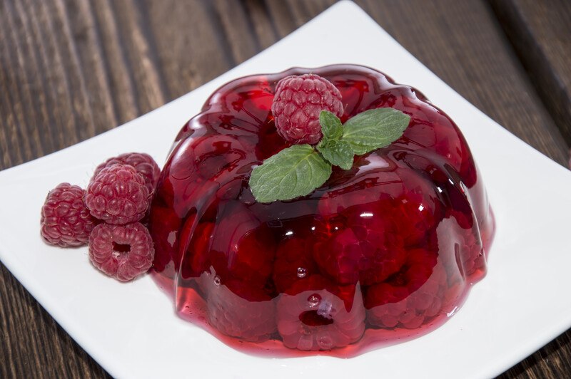 Red gelatin with raspberries on a white square plate rests on a wooden surface.