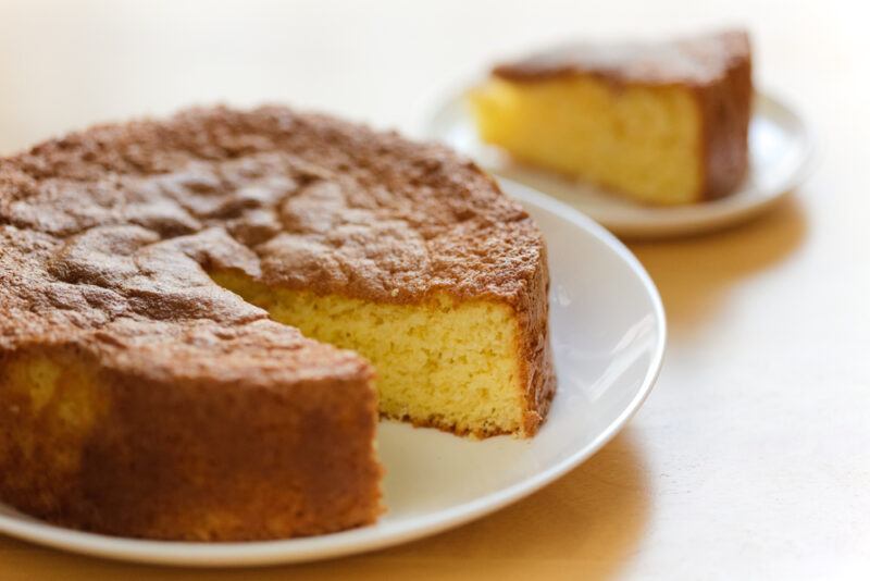 on a wooden surface is a white ceramic dish with a partially sliced Genoise cake, at the back is a small white ceramic dish with a slice of Genoise cake