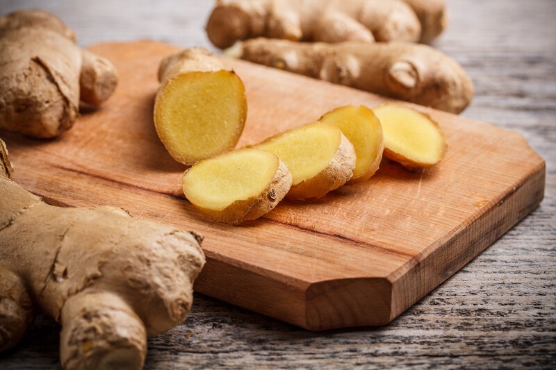 Five slices of ginger rest on a wooden cutting board next to several pieces of ginger root on a wooden surface.