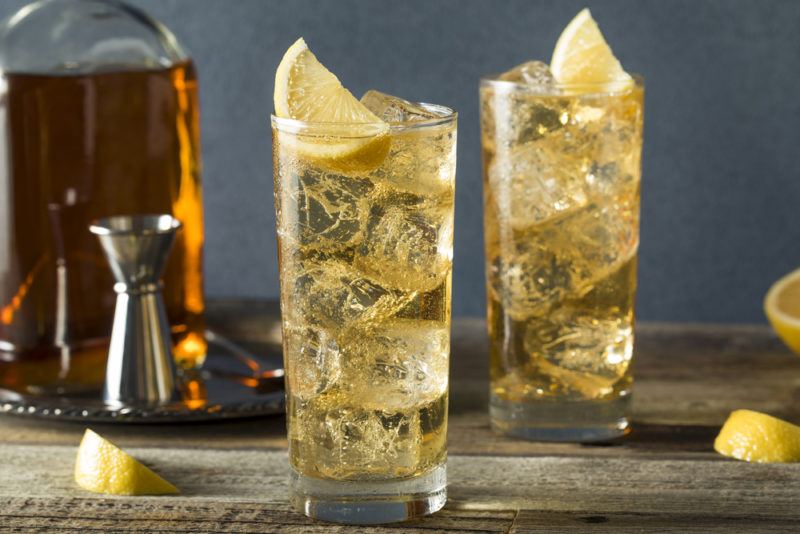 Two glasses of ginger ale with ice and lemon on a table, with a decanter and a jigger in the background