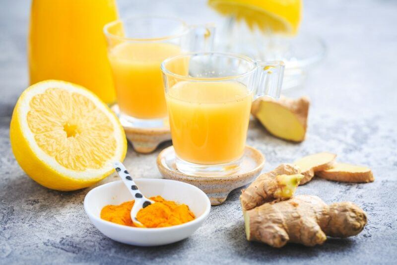 A gray table with ginger and turmeric shots, along with half a citrus fruit, turmeric powder, and ginger root