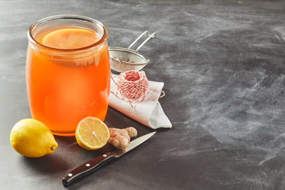 A glass jar of kombucha with a knife, lemon, and ginger on a table