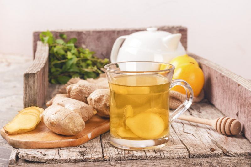 A glass mug of ginger tea with ginger, lemon and a tea pot in the background