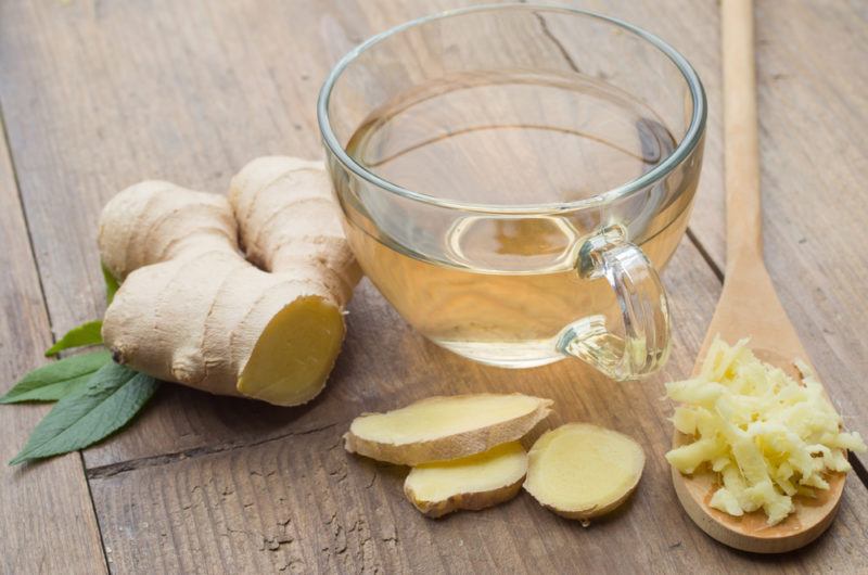 A glass mug containing ginger tea, with sliced and grated ginger on the table