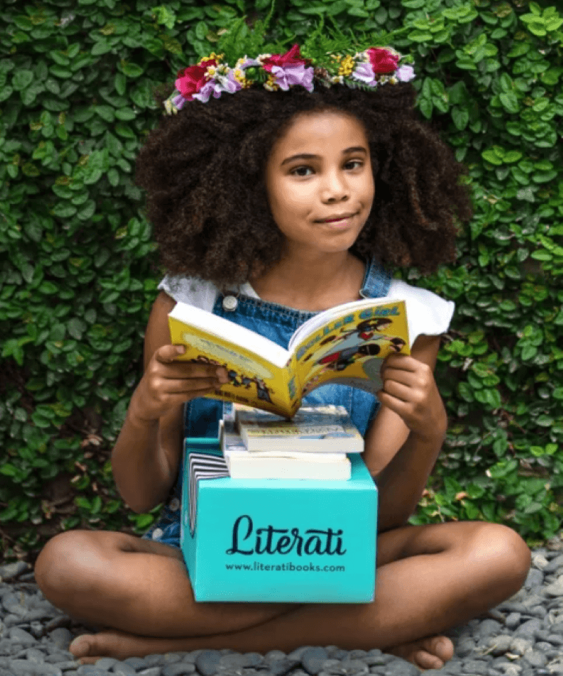 Girl sitting on the ground with a shrub behind reading a book with a Literati box and two books on top on her lap