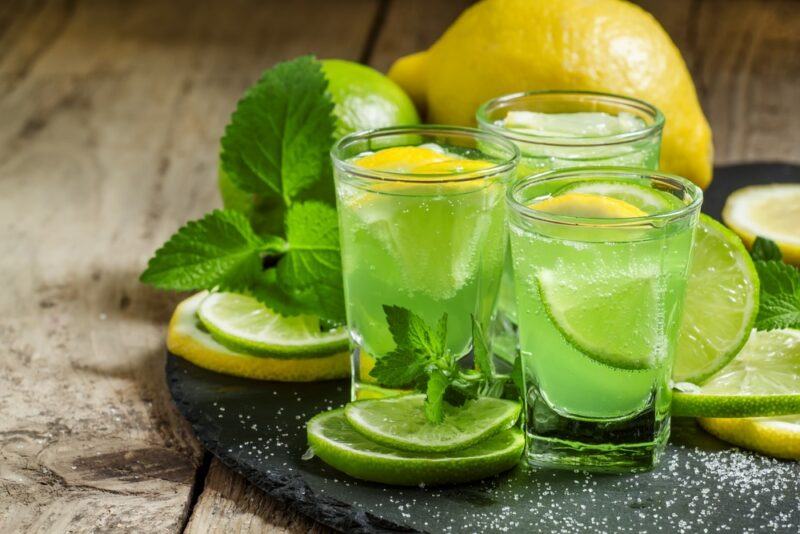 Three small glasses of a Patron Dew cocktail on a board in front of a lemon, a lime, and some mint leaves