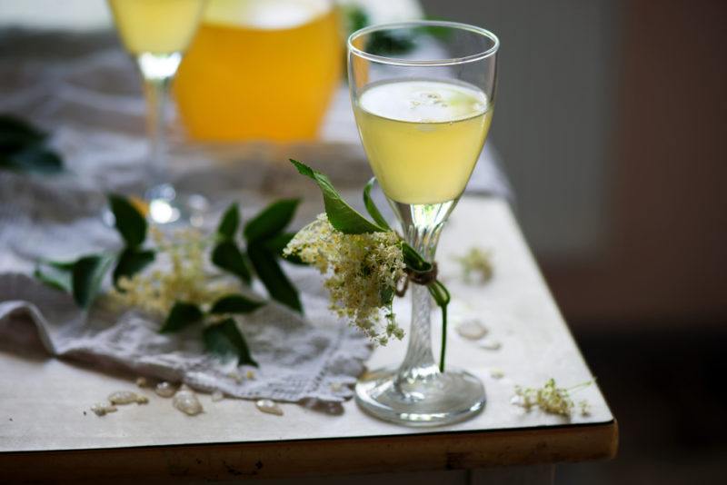 Two glasses of elderflower wine on a table, one in the foreground and one in the background, with elderflowers on the table