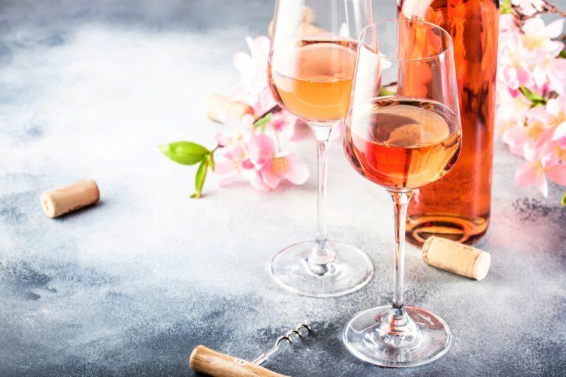 Two glasses of white zinfandel wine on a gray table, next to a bottle of the wine, some flowers, a cork, and a corkscrew