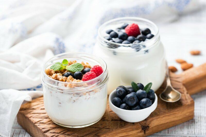 A wooden board with a large and a small glass jar of Greek yogurt with fruit and cereal on top, next to a small bowl of blueberries