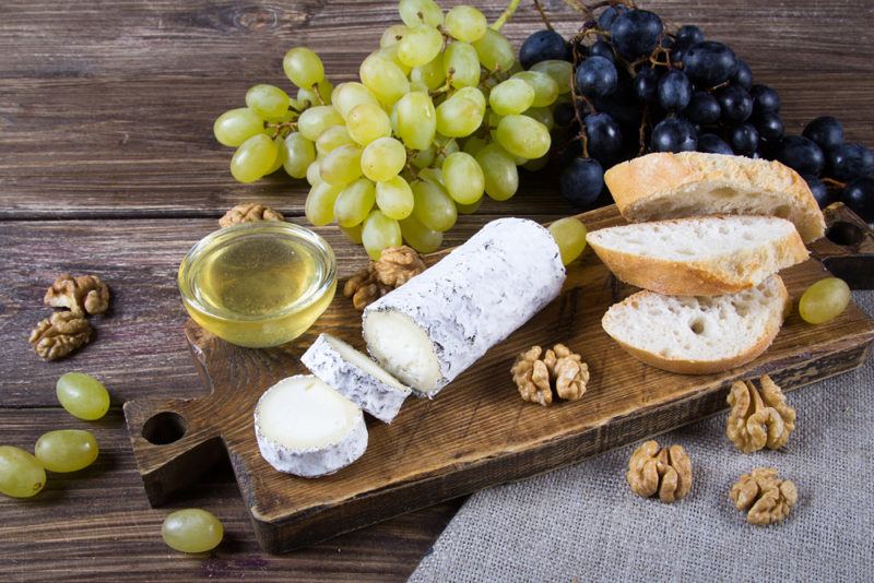 A wooden board with goats milk cheese, sliced bread, grapes and walnuts