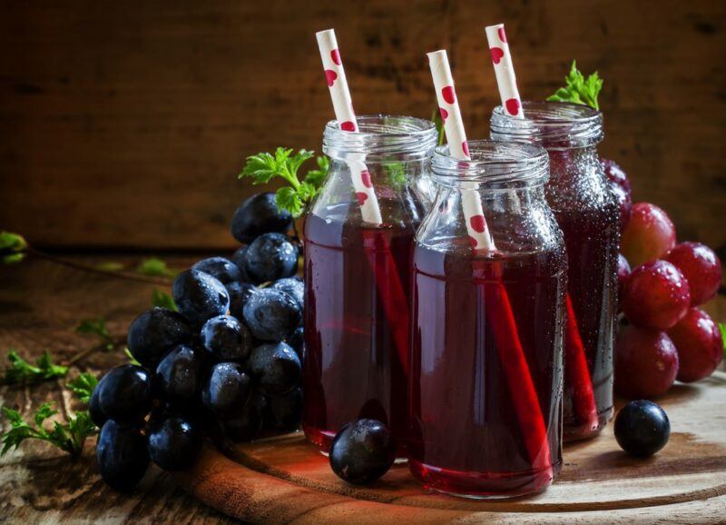 Three glass jars of grape juice with straws, with fresh grapes in the background