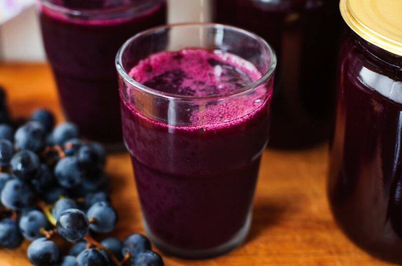 Two glasses of dark grape juice, next to jars of grape jam and some fresh grapes