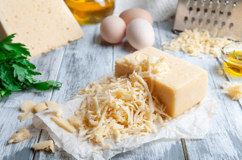 Grated semi hard cheese on paper, with a grater, eggs, oil and a wedge of the cheese in the background