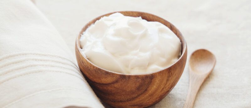 A wooden bowl filled with white Greek yogurt rests on a white cloth near a wooden spoon.