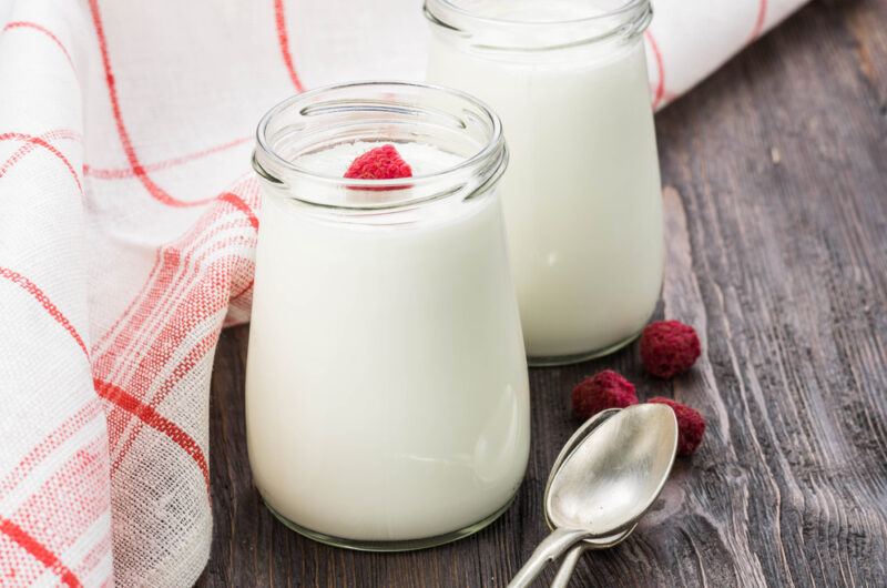 on a rustic wooden surface is a couple glasses of Greek yogurt with a raspberry on top, beside it is a metal spoons, loose raspberries, and a white table napkin with red stripes