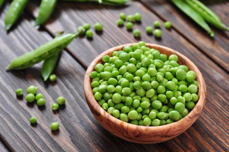 A wooden bowl of green peas and some green peas in their pod on a wooden table