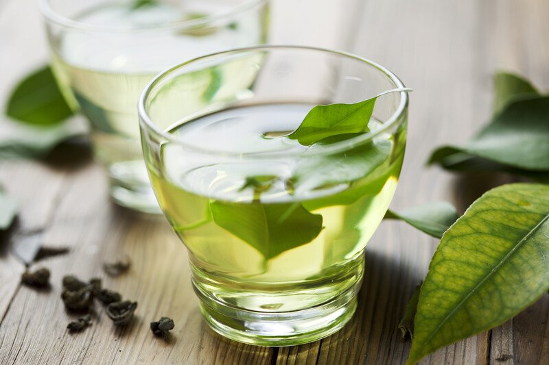 A clear glass with green tea and green leaves in it rests on a wooden table next to more green leaves and a second cup.