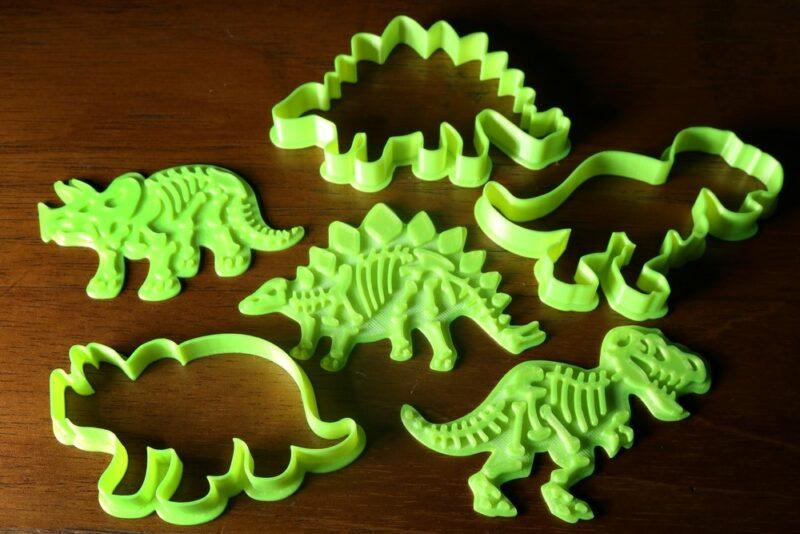 A wooden table with six neon green dinosaur cookie cutters. Three focus on outlines, while three provide the interior details