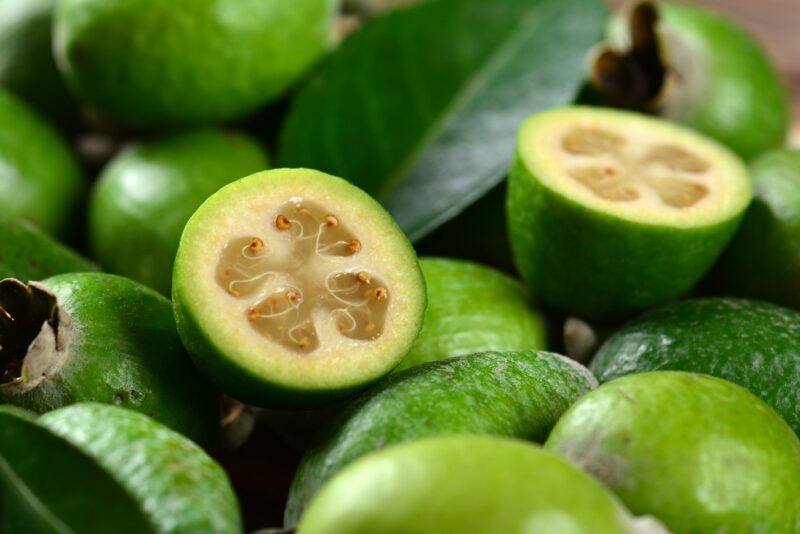 A pile of bright green feijoas with leaves, with two feijoa halves on top